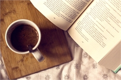 [ai] An overhead view of a white mug filled with coffee on a wooden cutting board next to an open book with text on its pages, placed on a patterned fabric.